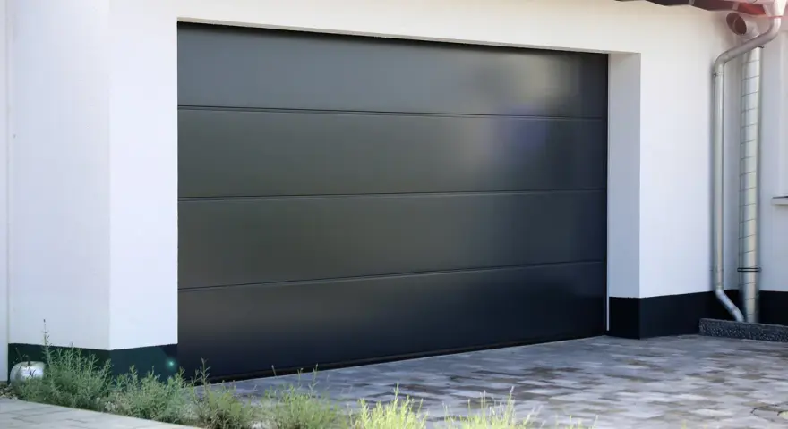 En modern, mörkgrå garageport montering med horisontella linjer. En vit fasad och grå stenläggning framför.