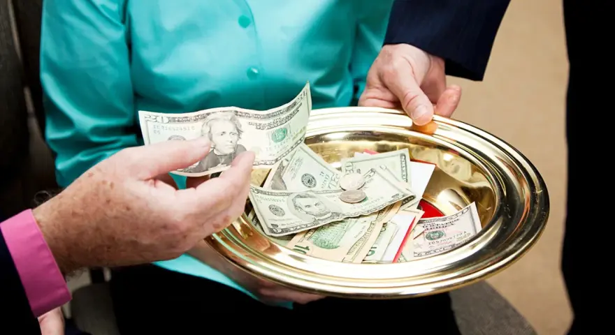 A hand places a twenty-dollar bill onto a golden tray filled with cash and coins, a gesture of offering to the poor.
