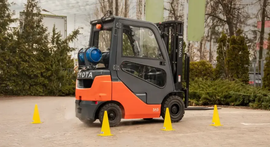 Pomarańczowo-szary wózek widłowy Toyota na placu manewrowym. Żółte pachołki wyznaczają trasę, sugerując kurs operatora wózka widłowego.