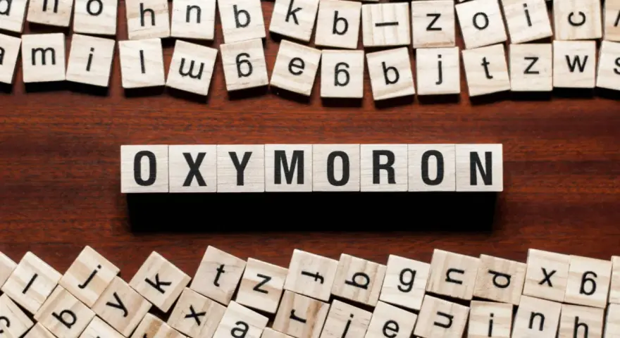 Holzbuchstaben bilden das Wort "OXYMORON". Sie sind umgeben von weiteren Buchstabenkacheln, die an Spiele wie Scrabble erinnern und Beispiele für Oxymoron-Begriffe darstellen könnten.