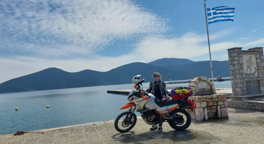 Kobieta z motocyklem nad morzem, z grecką flagą powiewającą na wietrze. Piękny dzień na podróż motocyklem nad morze.
