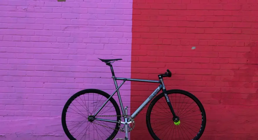 A sleek, fixed gear bike with "MASH" branding leans against a vibrant pink and red brick wall.