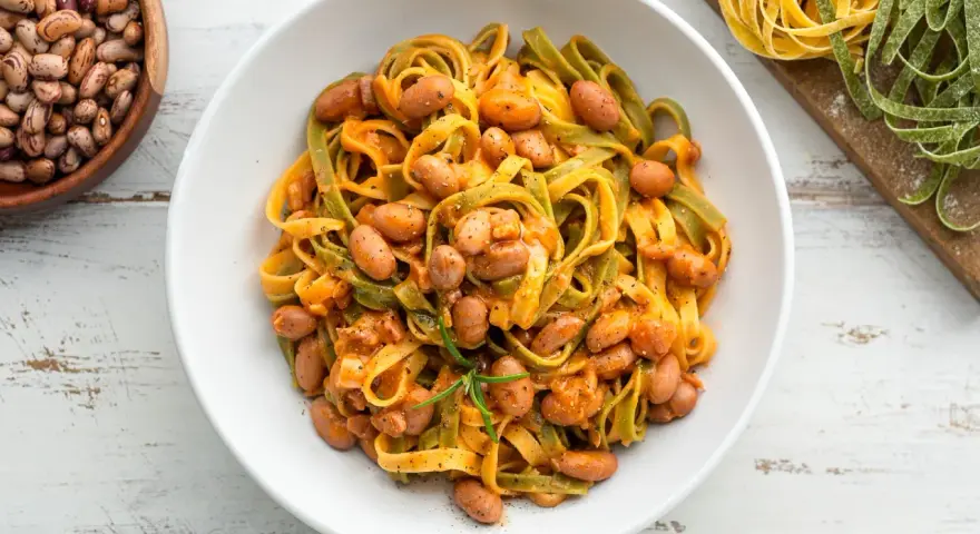 Pasta all'uovo con fagioli borlotti in una ciotola bianca, con fagioli secchi e pasta fresca colorata sullo sfondo.