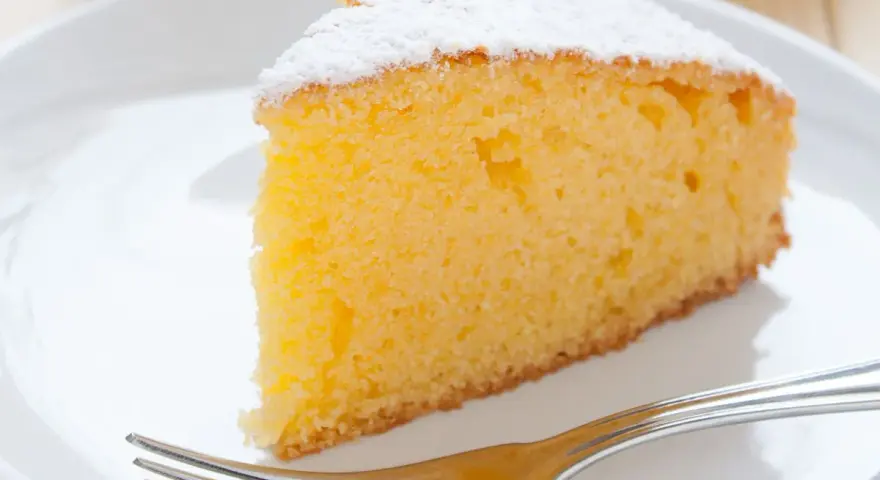 Ein saftiges Stück vanille kuchen, bestäubt mit Puderzucker, wartet auf einer weißen Platte mit einer Gabel.