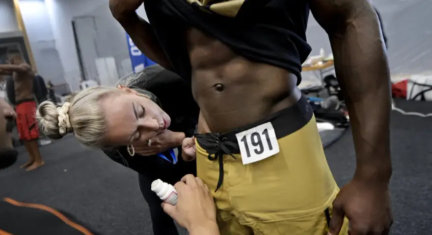 En kvinna applicerar olja på en man med en rippad kropp inför en tävling. Nummer 191 syns på hans shorts.