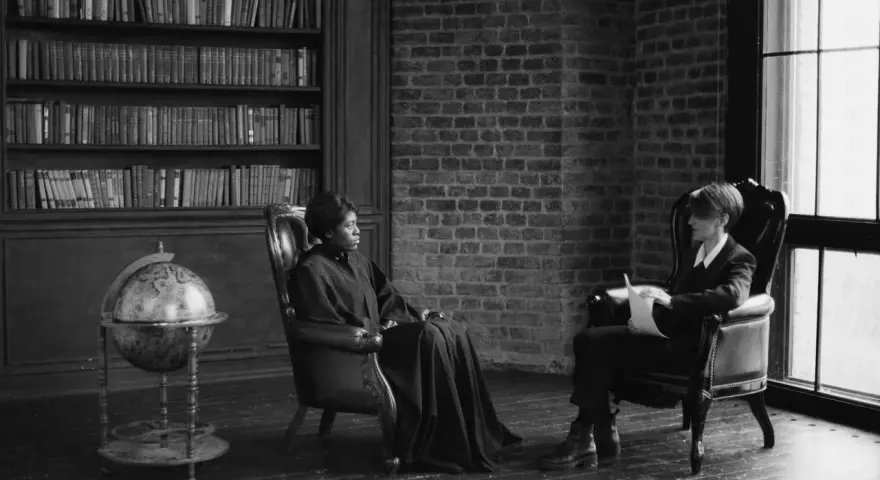 Deux personnes assises dans des fauteuils en cuir, une femme en robe longue et un homme en costume, discutent dans une bibliothèque. Scène évoquant des témoignages procédure en départage prud'hommes.