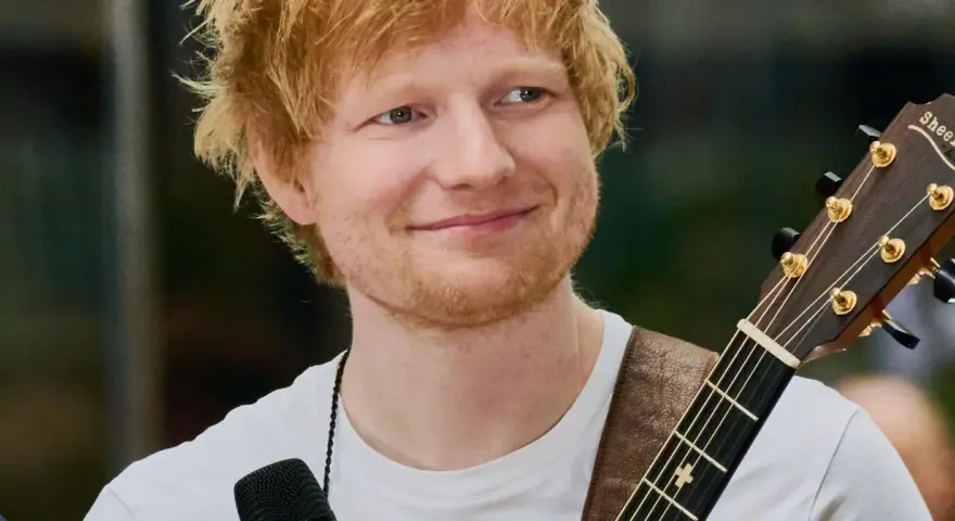 Ed Sheeran z gitarą, uśmiechnięty, w białej koszulce.