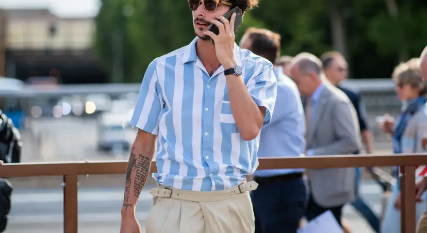 Hombre con camisa a rayas azules y blancas y tiro pantalón alto color beige, hablando por teléfono.
