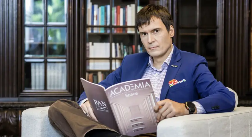 Mężczyzna w garniturze czyta magazyn "Academia Space" w bibliotece. W tle regały z książkami.