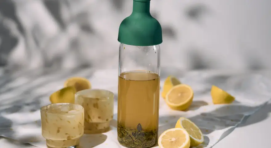 Orzeźwiający cold brew tea w butelce z zieloną zakrętką, obok szklanki z lodem i cytryny. Idealny na upalne dni.