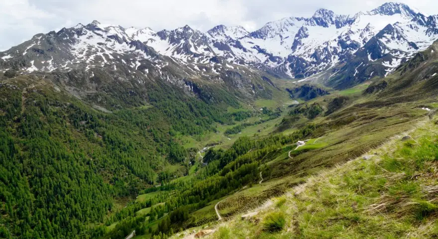 Un sentiero serpeggia su un pendio erboso verso un passo rombo in moto, con montagne innevate sullo sfondo.