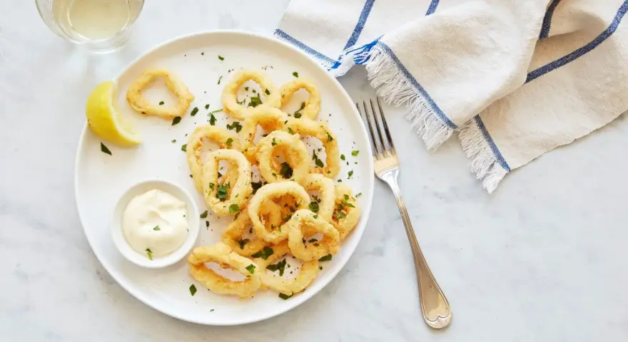 Crujientes calamares a la romana, servidos con limón y salsa tártara en un plato blanco.