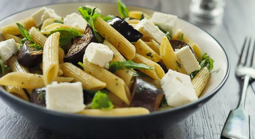 Piatto di pasta con melanzane in bianco, cubetti di formaggio fresco e foglie verdi.