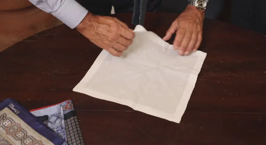 Mani esperte mostrano come piegare fazzoletto da taschino su un tavolo di legno. Altri fazzoletti colorati sono visibili a sinistra.