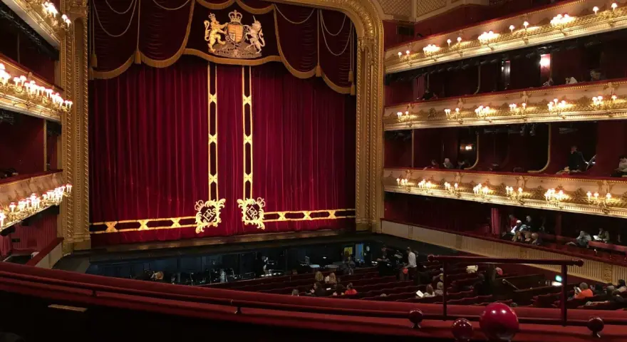 Ein prunkvolles Opernhaus mit roten Samtvorhängen und goldenen Verzierungen. Die Logen sind mit Lichtern geschmückt, und im Parkett sitzen Zuschauer.