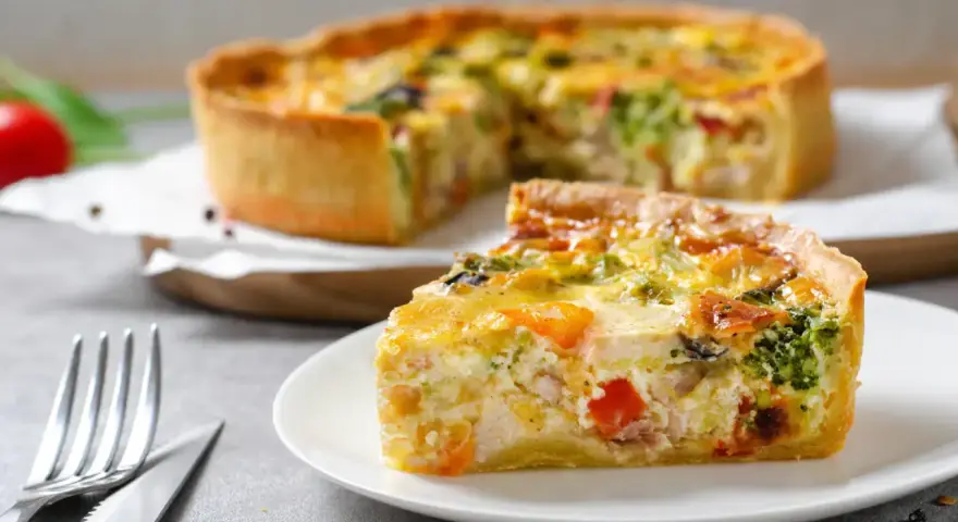 Une part de quiche savoureuse avec une pâte à tarte maison dorée, garnie de légumes et de poulet. Le reste de la tarte est flou en arrière-plan.
