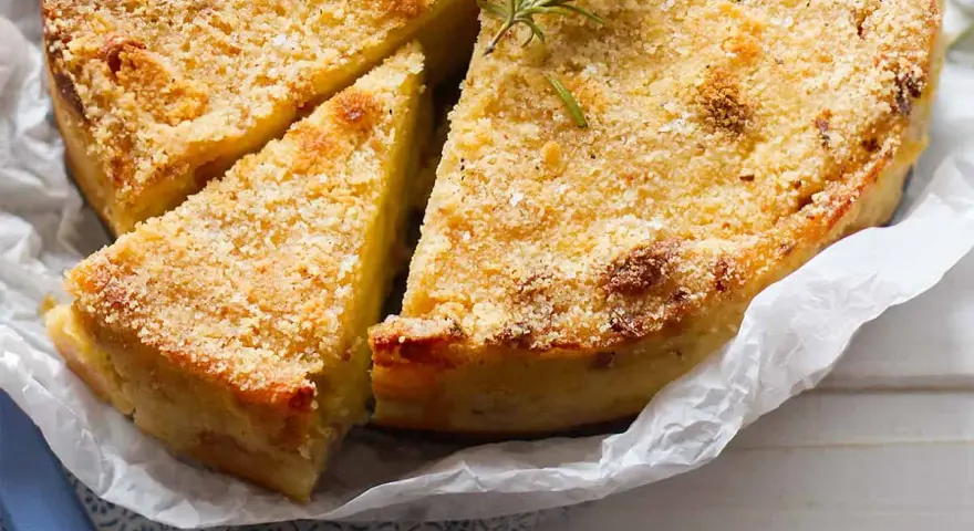 Delizioso gateau di patate dorato e croccante, guarnito con rosmarino fresco. Una fetta è già stata tagliata, invitando ad assaggiarlo.