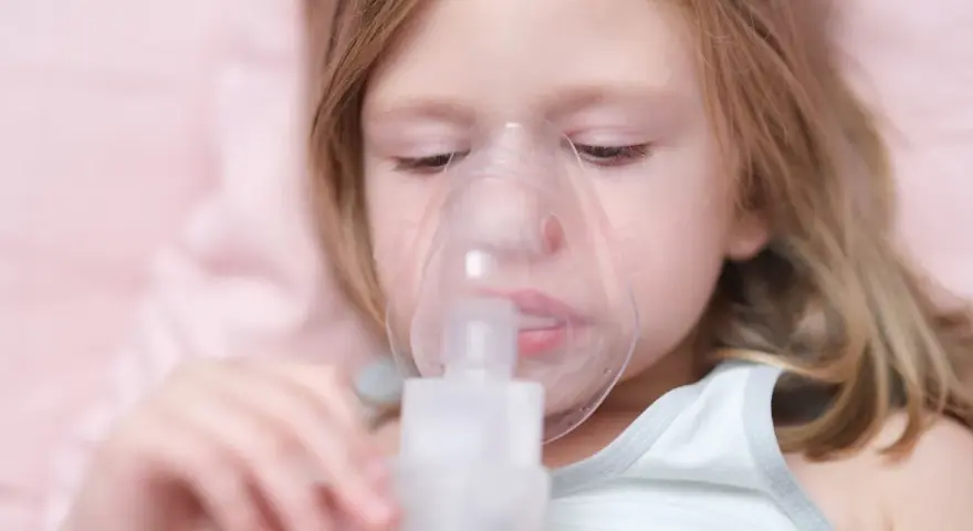 Mała dziewczynka z maską inhalacyjną na twarzy. Dziecko przechodzi inhalację na kaszel, by poczuć ulgę.