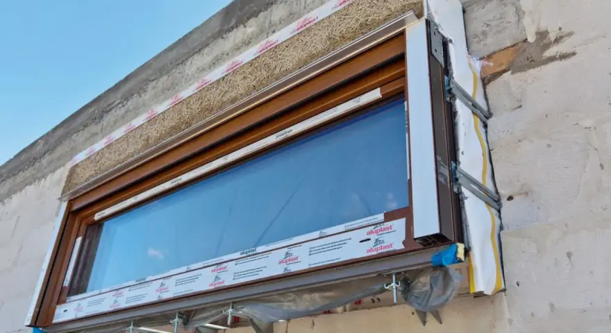 Ocieplenie okien od wewnątrz z użyciem naturalnych materiałów izolacyjnych, widoczne na tle betonowej ściany i błękitnego nieba.