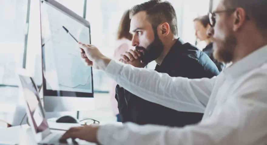 Analityk biznesowy wskazuje na wykres na monitorze, omawiając dane z kolegą.