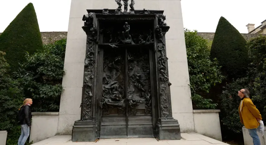La Porte de l'Enfer de Rodin, une œuvre monumentale en bronze, se dresse au milieu d'un jardin. Deux visiteurs contemplent cette sculpture complexe.
