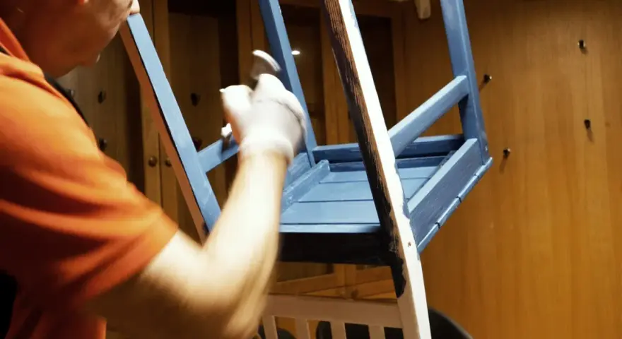Person i orange tröja målar en blå stol med pensel.