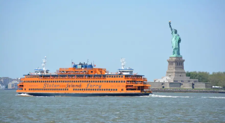 Pomarańczowy prom Staten Island Ferry płynie obok Statuy Wolności.