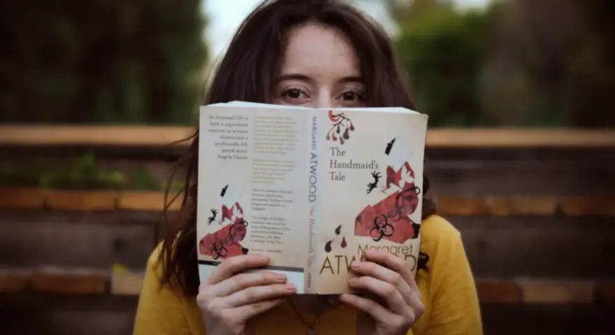 Młoda kobieta w żółtej bluzce czyta "Opowieść podręcznej" Margaret Atwood. Jej oczy błyszczą, gdy zagłębia się w świat literatury, co jest idealnym przykładem pasji do filologii.