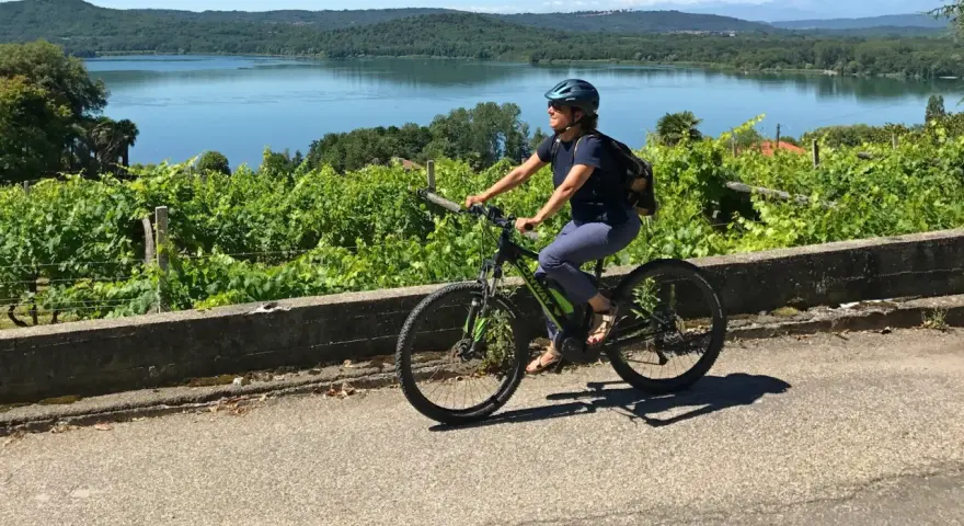 Donna in bici elettrica lungo il cammino di Oropa, tra vigneti e un lago sullo sfondo.