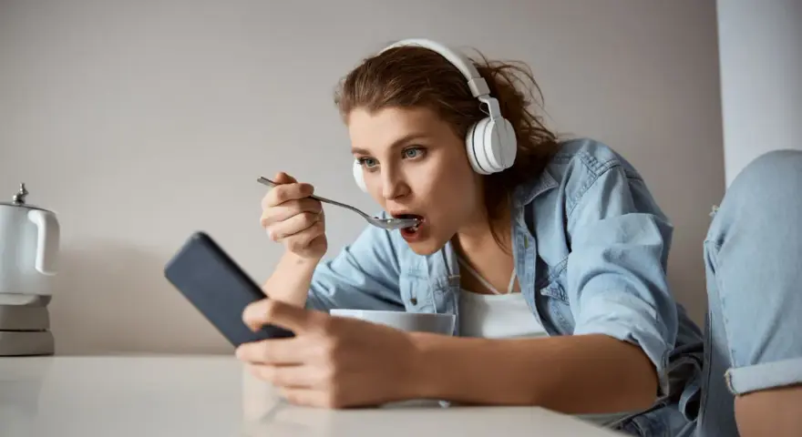 Młoda kobieta w słuchawkach je płatki, patrząc w telefon. Czy to objawy fonoholizmu?