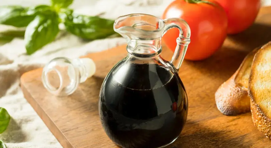 Mały dzbanuszek z ciemnym, gęstym **octem balsamicznym** obok pomidorów, bazylii i grzanek.