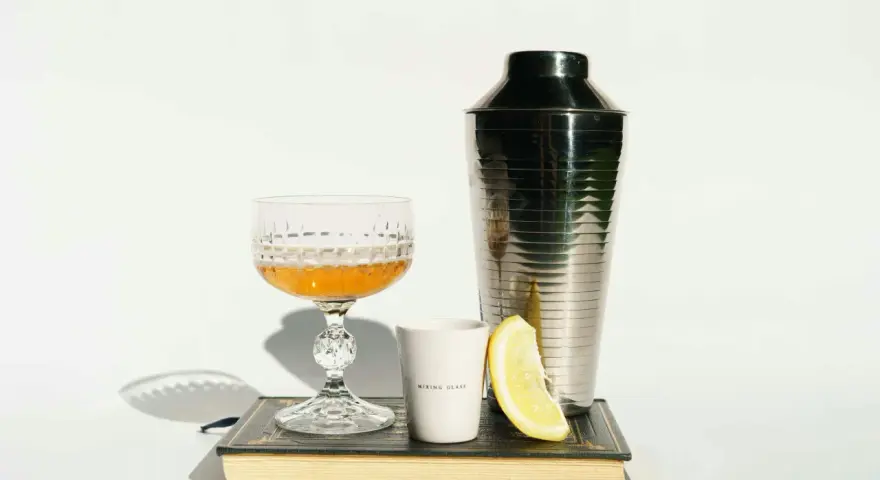 Elegancki kieliszek z bursztynowym trunkiem, shaker, plasterek cytryny i biały kubeczek z napisem "MIXING GLASS" – wszystko to sugeruje, z czym pić whisky, by stworzyć wyrafinowany koktajl.
