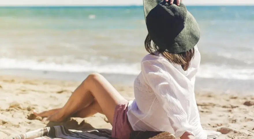 Kobieta w kapeluszu siedzi na plaży, ciesząc się spokojnymi miejscami nad morzem. Słońce i szum fal tworzą idealną atmosferę relaksu.