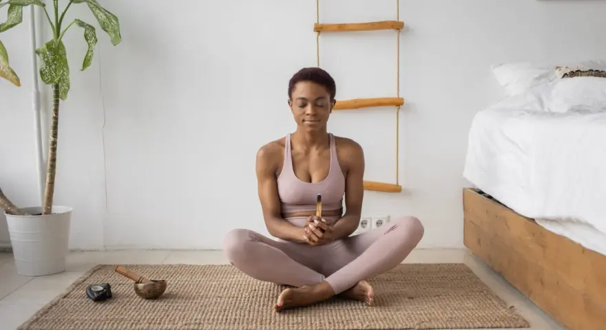 Mujer meditando en postura de loto, con los ojos cerrados y un palo de incienso en las manos. A su lado, un cuenco tibetano y una vela. Aprende como meditar para encontrar la paz interior.