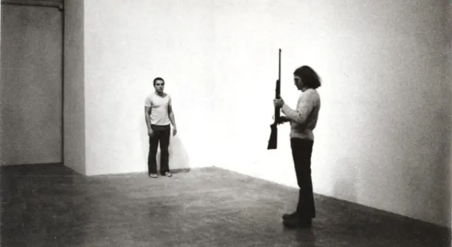Chris Burden, dans une performance tendue, tient un fusil. Un homme se tient à distance, le regard fixé.