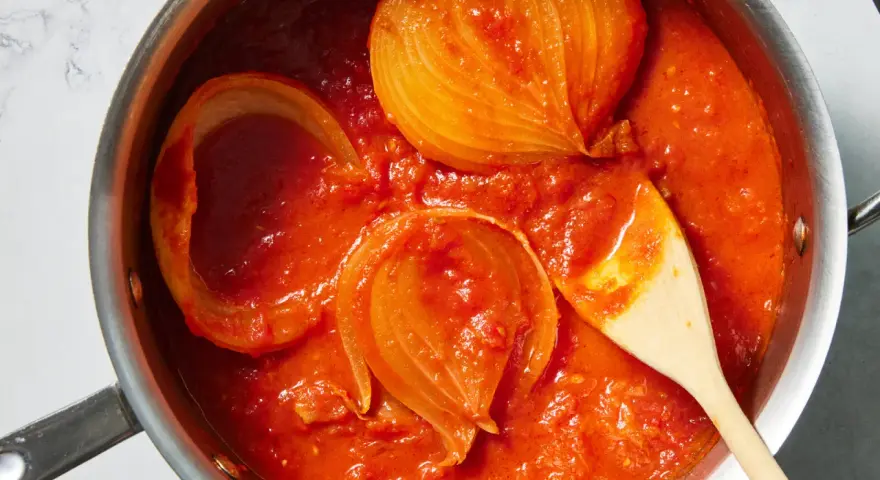 Tomatensoße mit Zwiebelstücken in einem Topf, bereit für Marcella Hazan Rezepte. Ein Holzlöffel rührt die köstliche Soße.