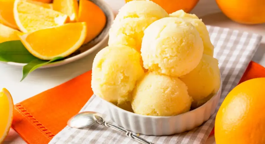 Erfrischendes orangen sorbet in einer Schale, umgeben von frischen Orangen und saftigen Orangenstücken. Ein Löffel liegt bereit.