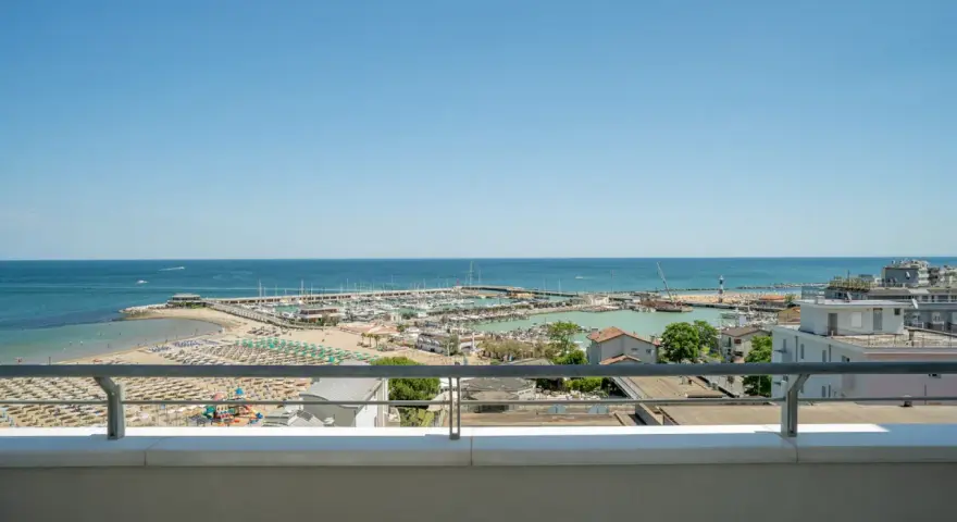 Vista dal balcone dell'hotel Acropolis Cattolica: spiaggia, porto turistico e mare azzurro sotto un cielo limpido.