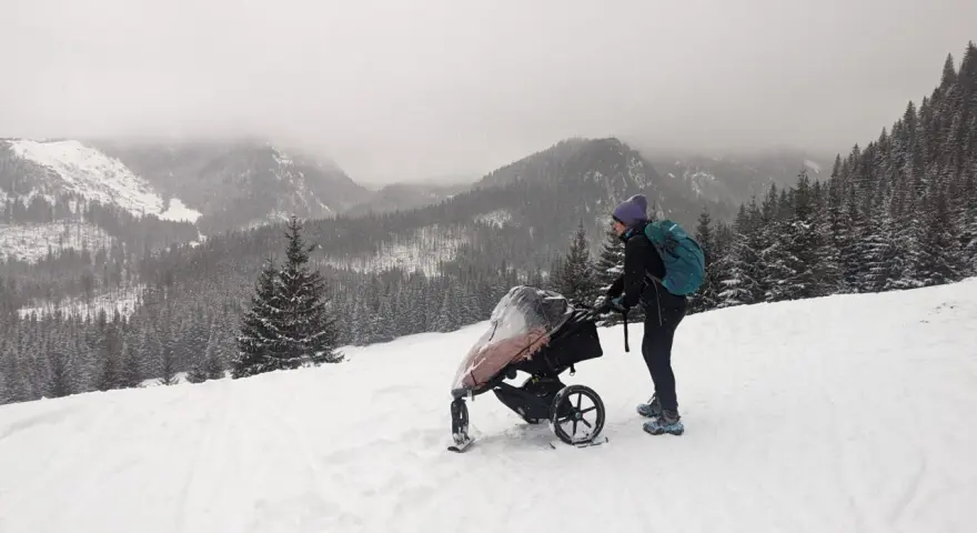 Zimowe Tatry z wózkiem. Kobieta w czapce i z plecakiem pcha wózek po ośnieżonym stoku, podziwiając górski krajobraz.