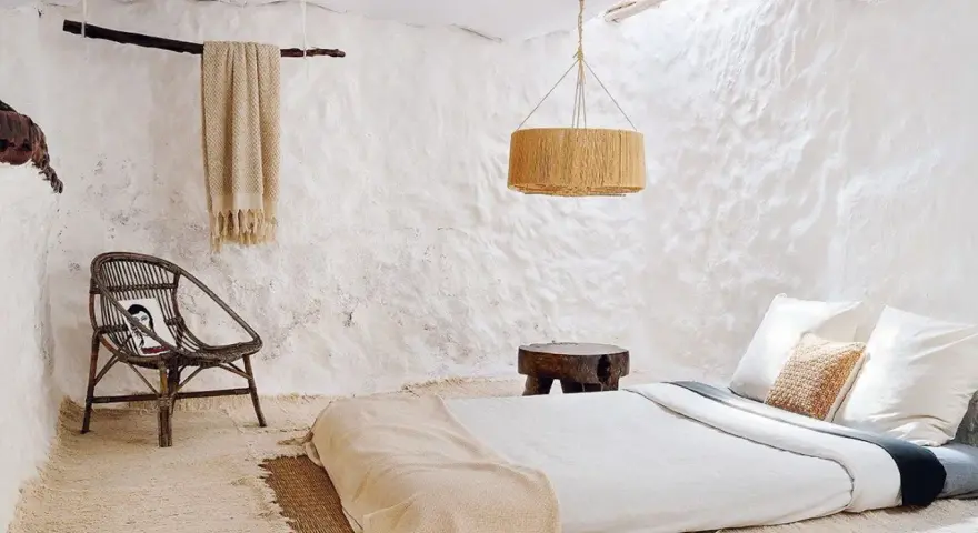 Habitación rústica con paredes de encalar, cama baja, silla de mimbre y manta de lana.