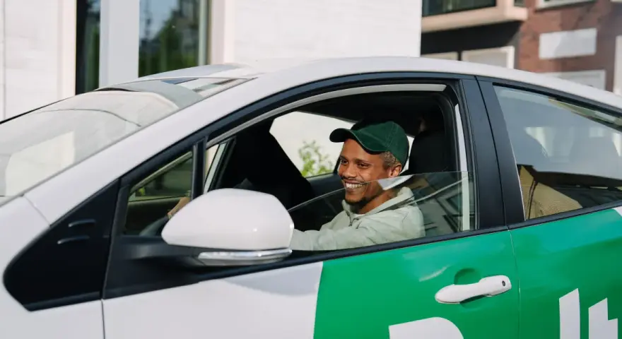 Lächelnder Mann mit grüner Kappe am Steuer eines weißen Autos mit grünen Bolt-Aufklebern. Er wird bald ein Bolt-Fahrer sein.