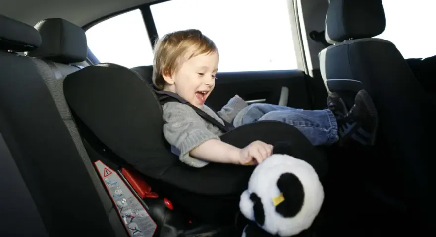 Ein fröhliches Kind sitzt im Kindersitz im Auto und spielt mit einem Panda-Stofftier.