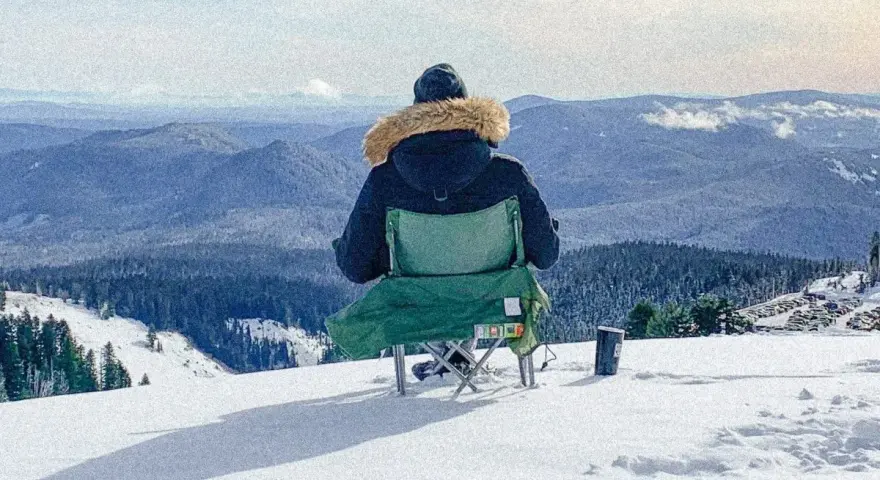 Persona sentada en silla plegable en la nieve, mirando un paisaje montañoso cubierto de árboles.