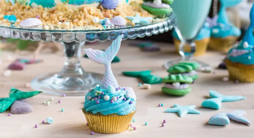 Meerjungfrau Muffins mit blauer Glasur und Schwanz-Dekorationen, umgeben von Muscheln und Seesternen.