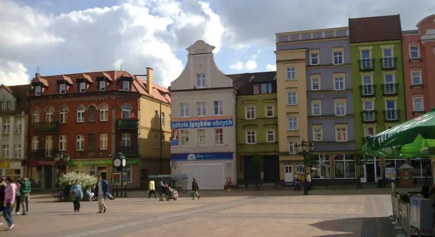 Rynek w Chojnicach z kolorowymi kamienicami, w tym ratusz. Ludzie spacerują po brukowanym placu.