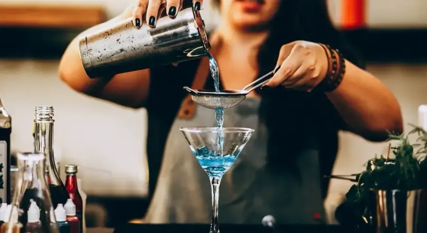 Barman przygotowuje drinka, przelewając niebieski płyn przez sitko do kieliszka. W tle widać butelki z różnymi nazwami drinków alkoholowych.