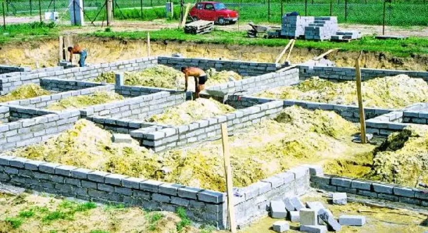 Budowa fundamentu z bloczków betonowych. Dwóch robotników pracuje przy wznoszeniu ścian fundamentowych.