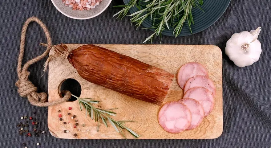 Kiełbasa żywiecka na desce, obok gałązka rozmarynu, czosnek i kolorowy pieprz. Kilka plastrów już pokrojonych.