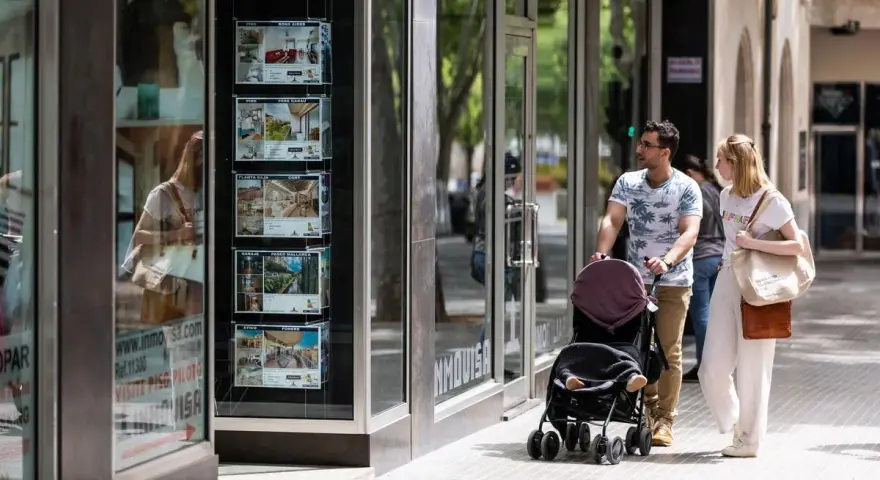 Pareja pasea con carrito, mirando escaparate inmobiliario. Quizás planean donar un piso a un hijo en Cataluña.