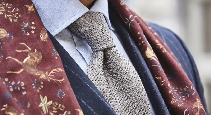 Elegancki mężczyzna w błękitnej koszuli, szarym krawacie i bordowym szalu z motywem jeleni. Całość emanuje nonszalancką elegancją, czyli sprezzaturą.